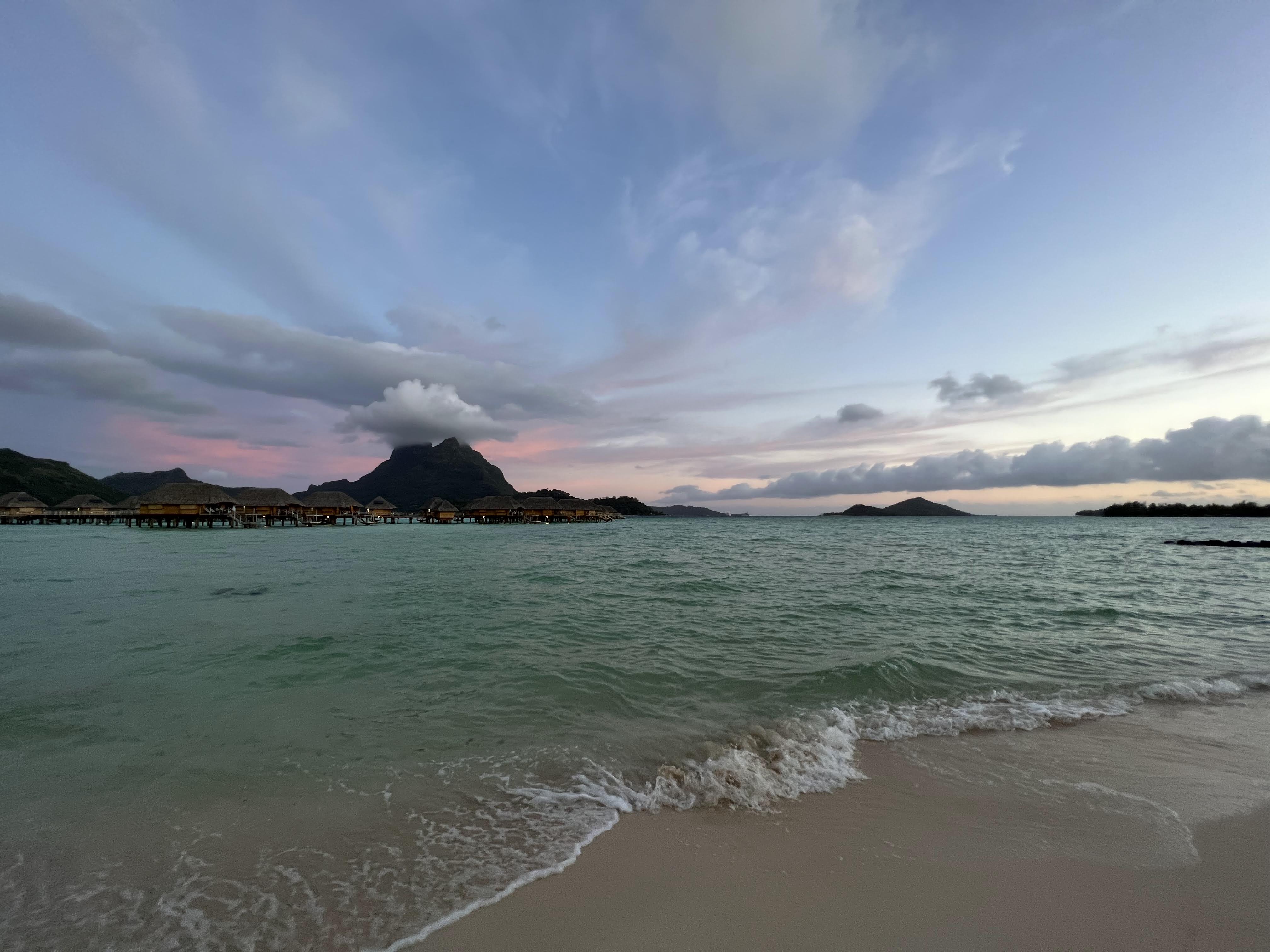 Tropical paradise with overwater bungalows on a pristine beach, turquoise ocean waters, and lush green mountains in the background during golden hour