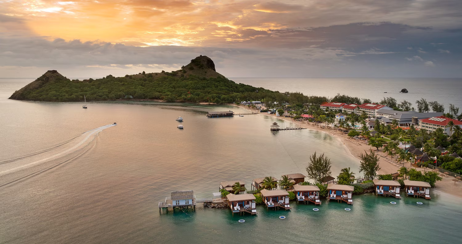Sandals St. Lucian resort featuring overwater bungalows with thatched roofs, pristine beach, turquoise bay waters, and lush green mountains in the background during golden hour